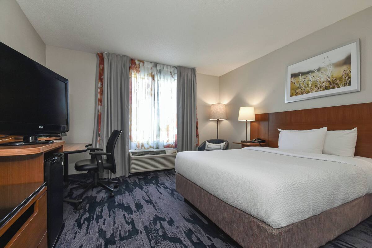 A hotel room with a large bed, a desk with a flat-screen TV, a chair, a lamp, and a painting on the wall.