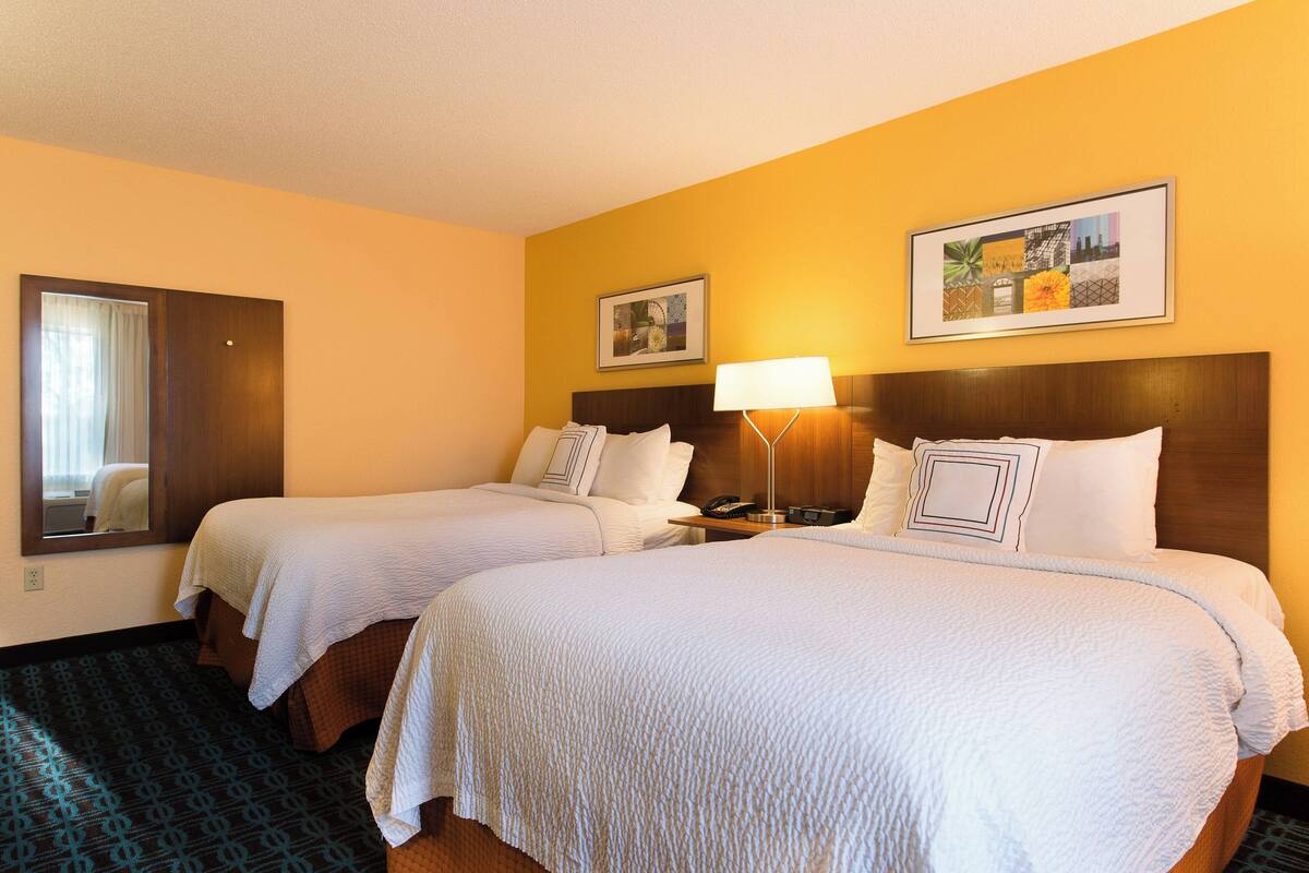 A hotel room with two beds, a bedside table with a lamp, a mirror, and two framed pictures on the wall.