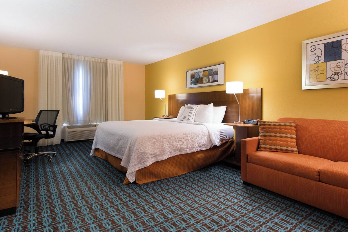 A hotel room with a large bed, a sofa, a desk with a chair, and a TV.