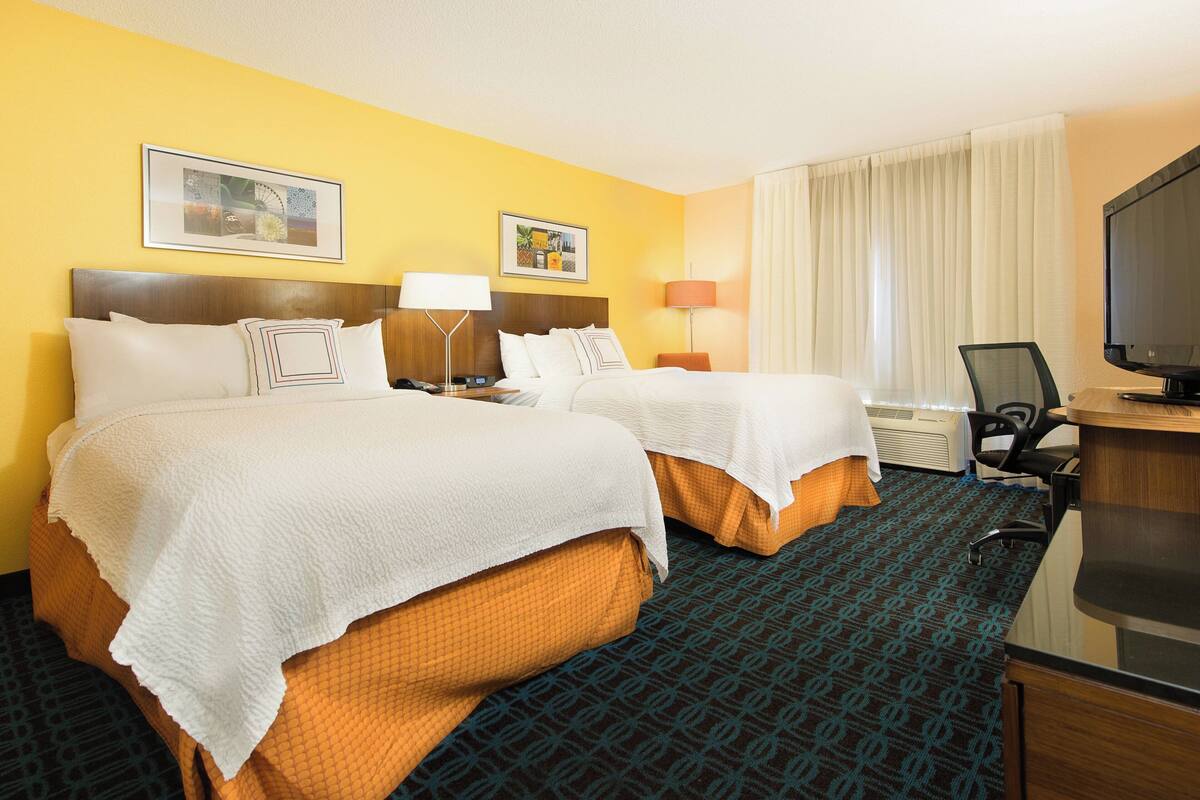 A hotel room with two beds, a desk, a television, and a window with curtains.