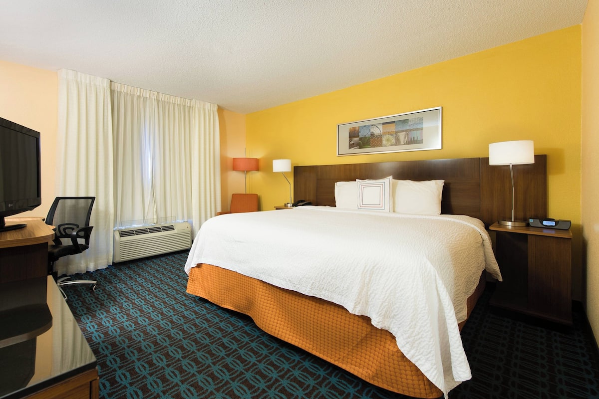 A hotel room with a bed, a desk with a computer, an air conditioner, a lamp, and a painting on the wall.