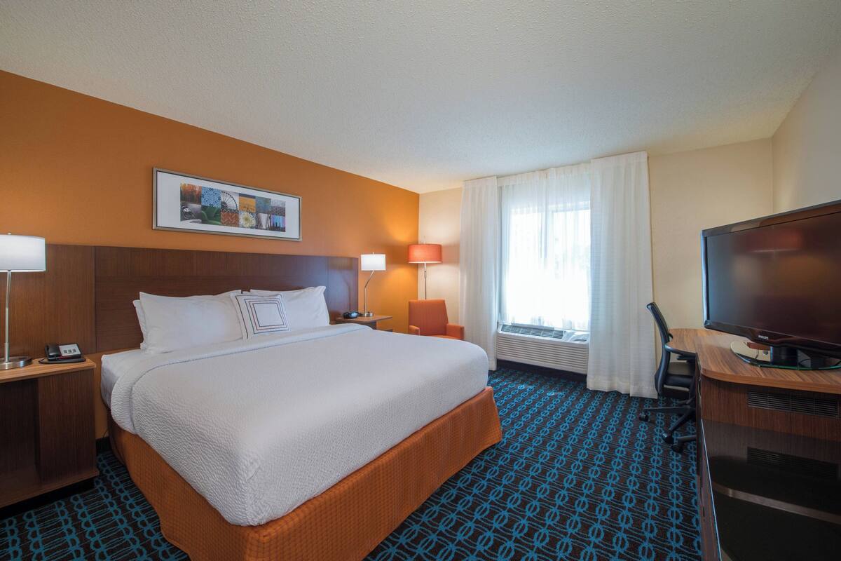A hotel room with a large bed, a desk with a flat-screen TV, a chair, a desk lamp, and a framed picture on the wall.