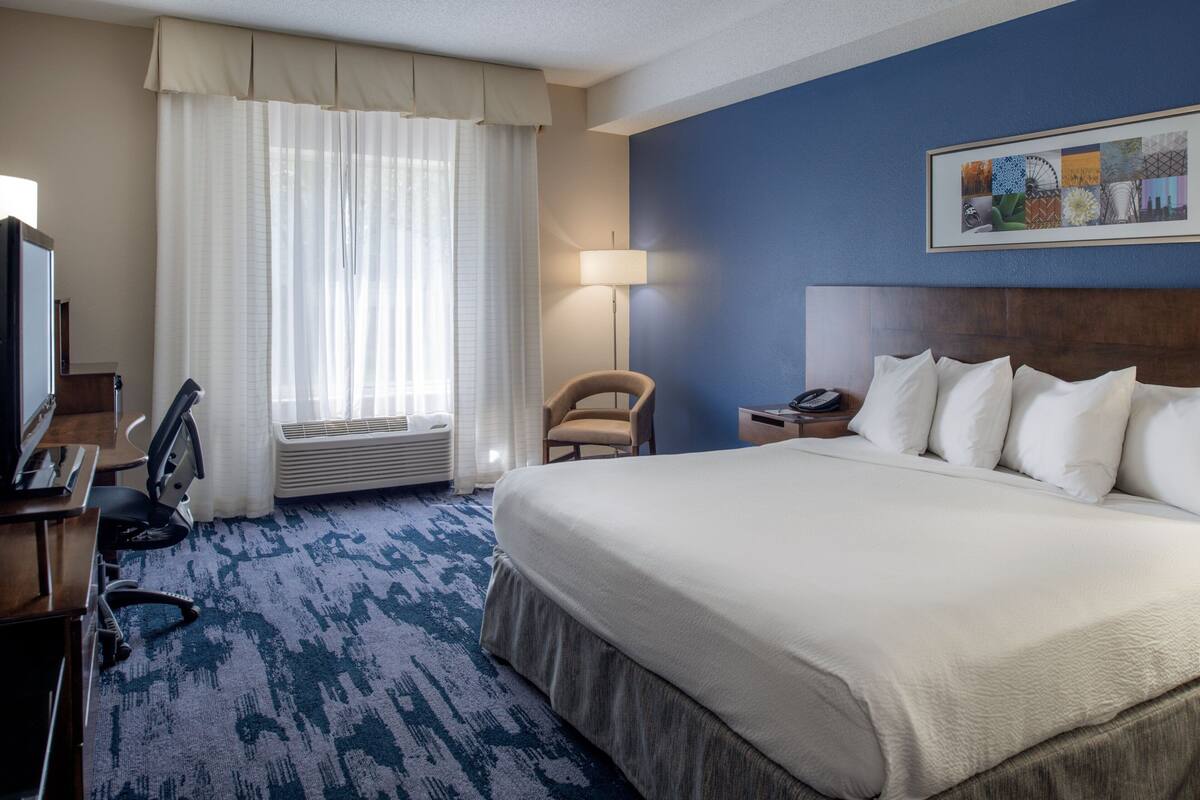A hotel room with a large bed, a desk with a chair, a lamp, and a window with curtains.