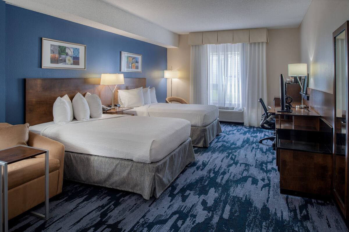 A hotel room with a large bed, a smaller bed, a desk, a chair, and a desk lamp.