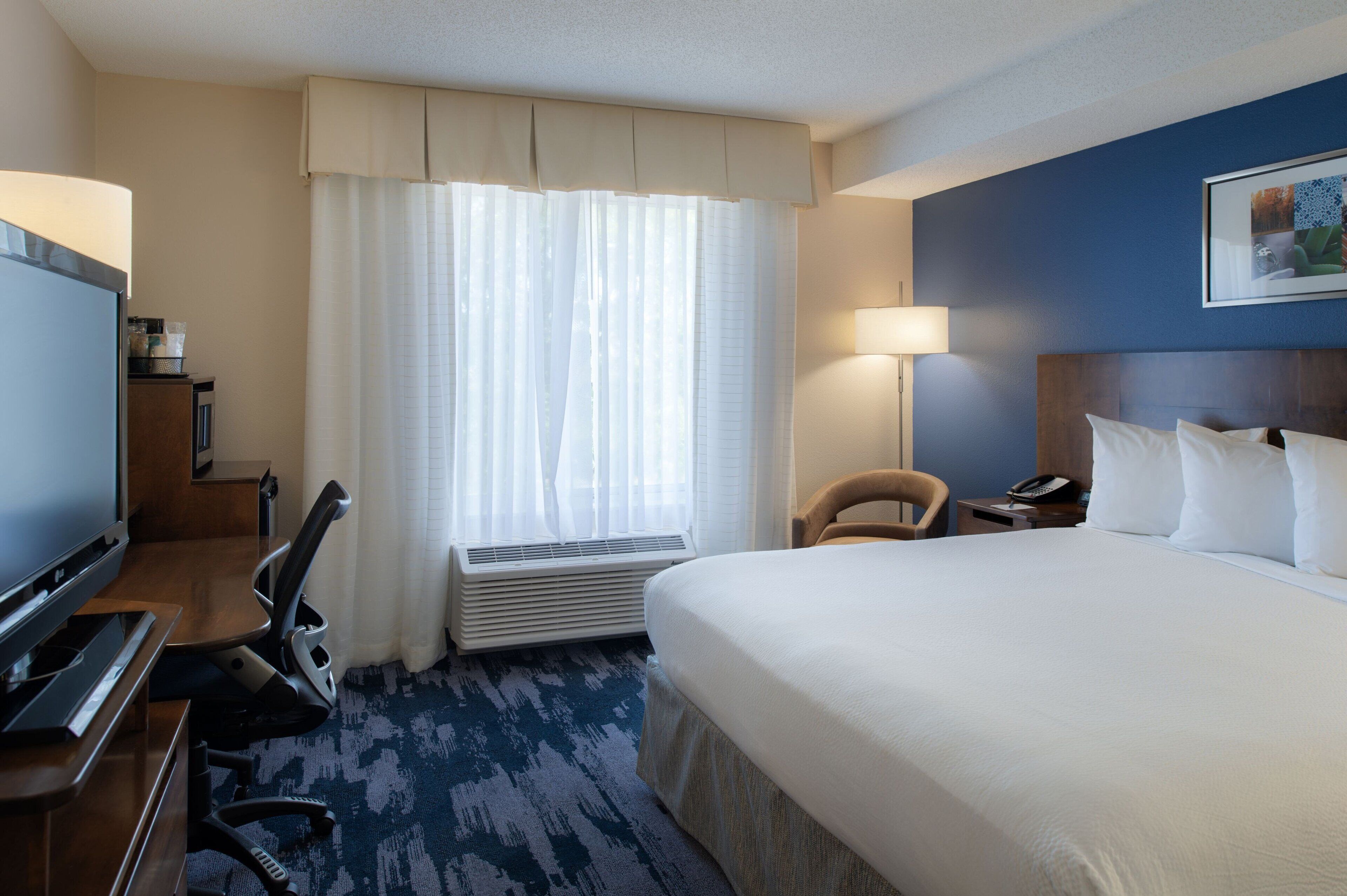 A hotel room with a large bed, a desk with a flat-screen TV, a chair, a lamp, and a window with curtains.