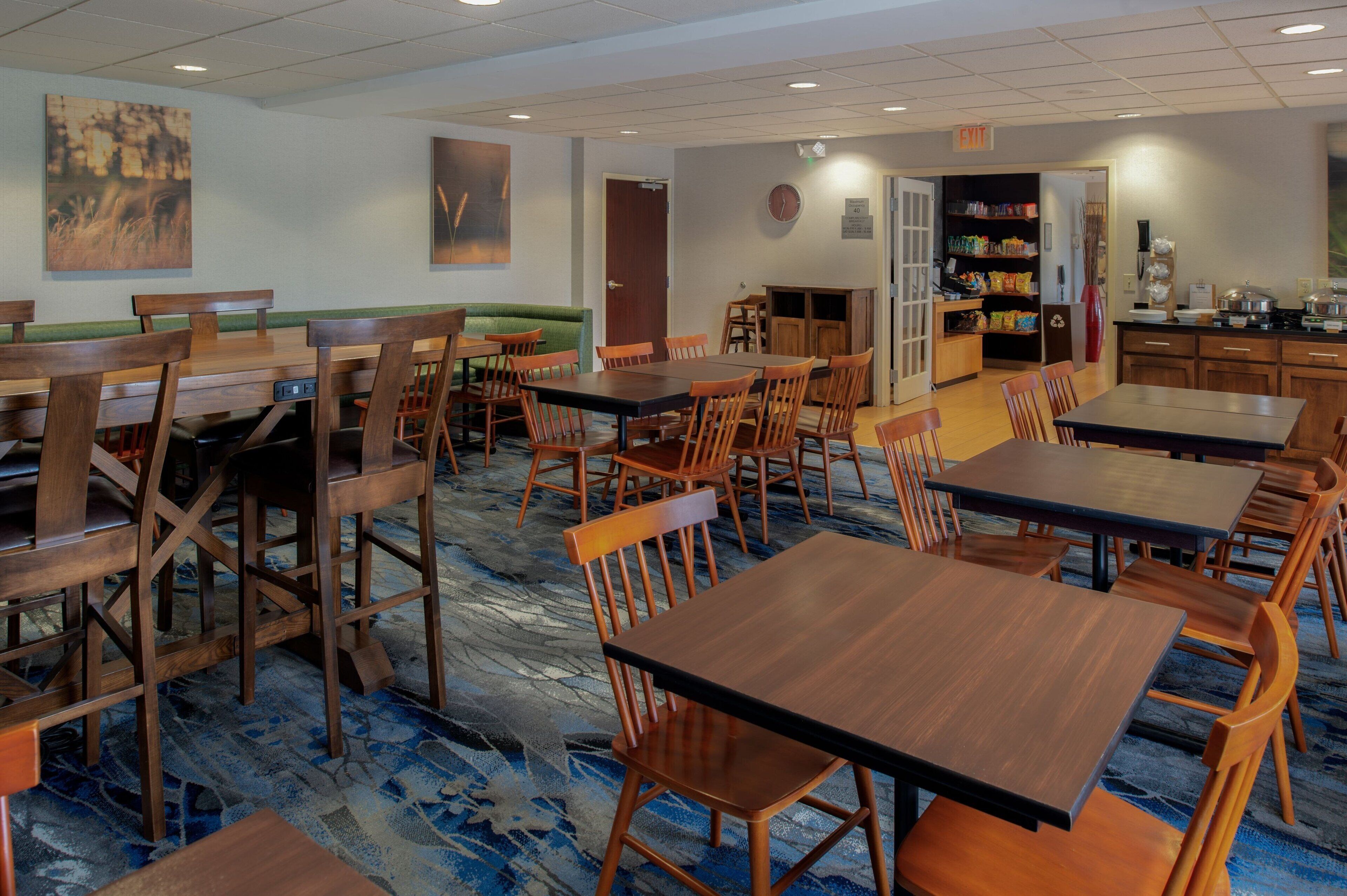 A dining area with multiple wooden tables and chairs, a green cushioned bench, and a kitchen area with shelves stocked with food items.