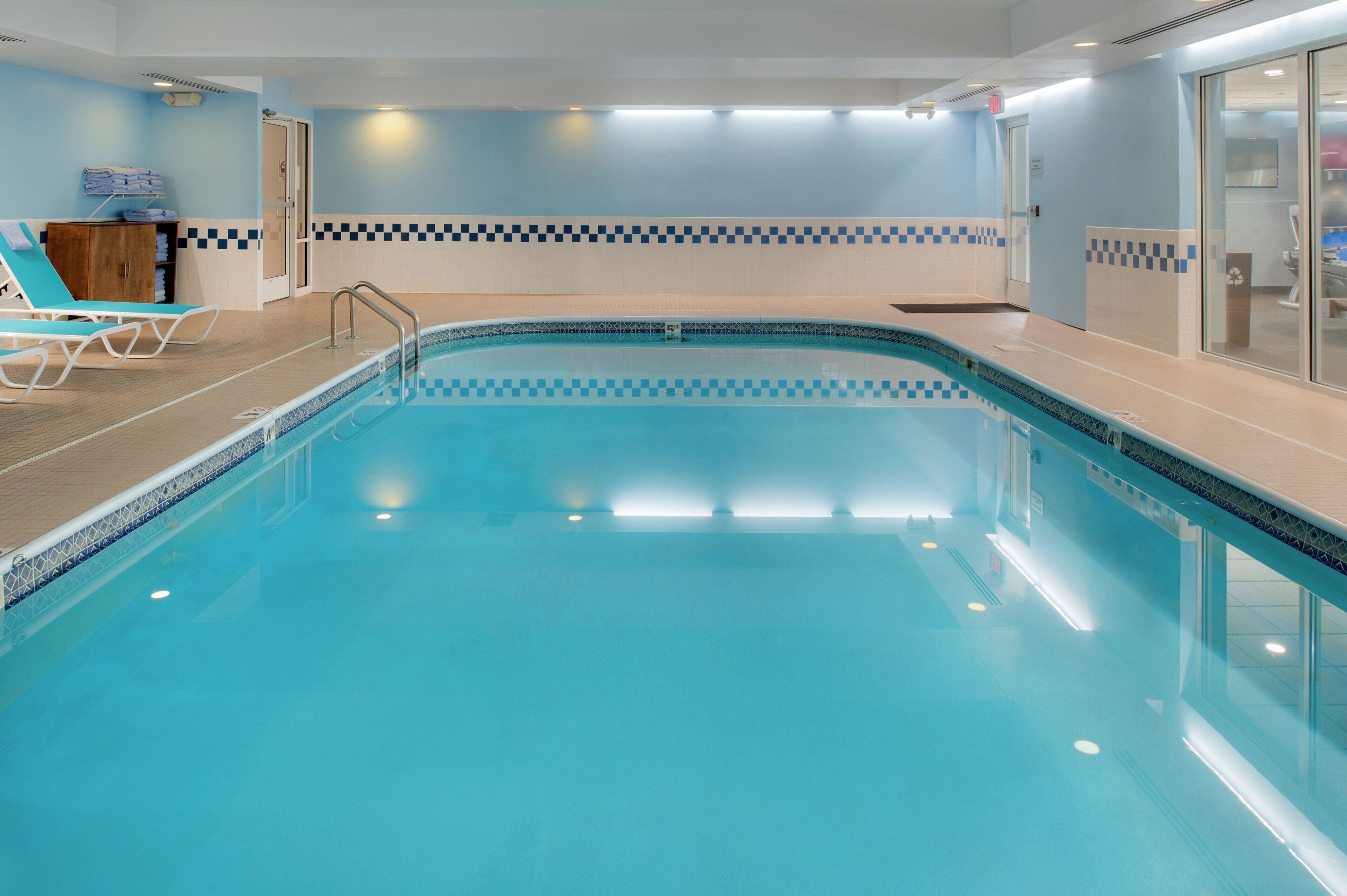 Una piscina interior con borde de azulejos, zona de madera con terrazza y algunas sillas de descanso.