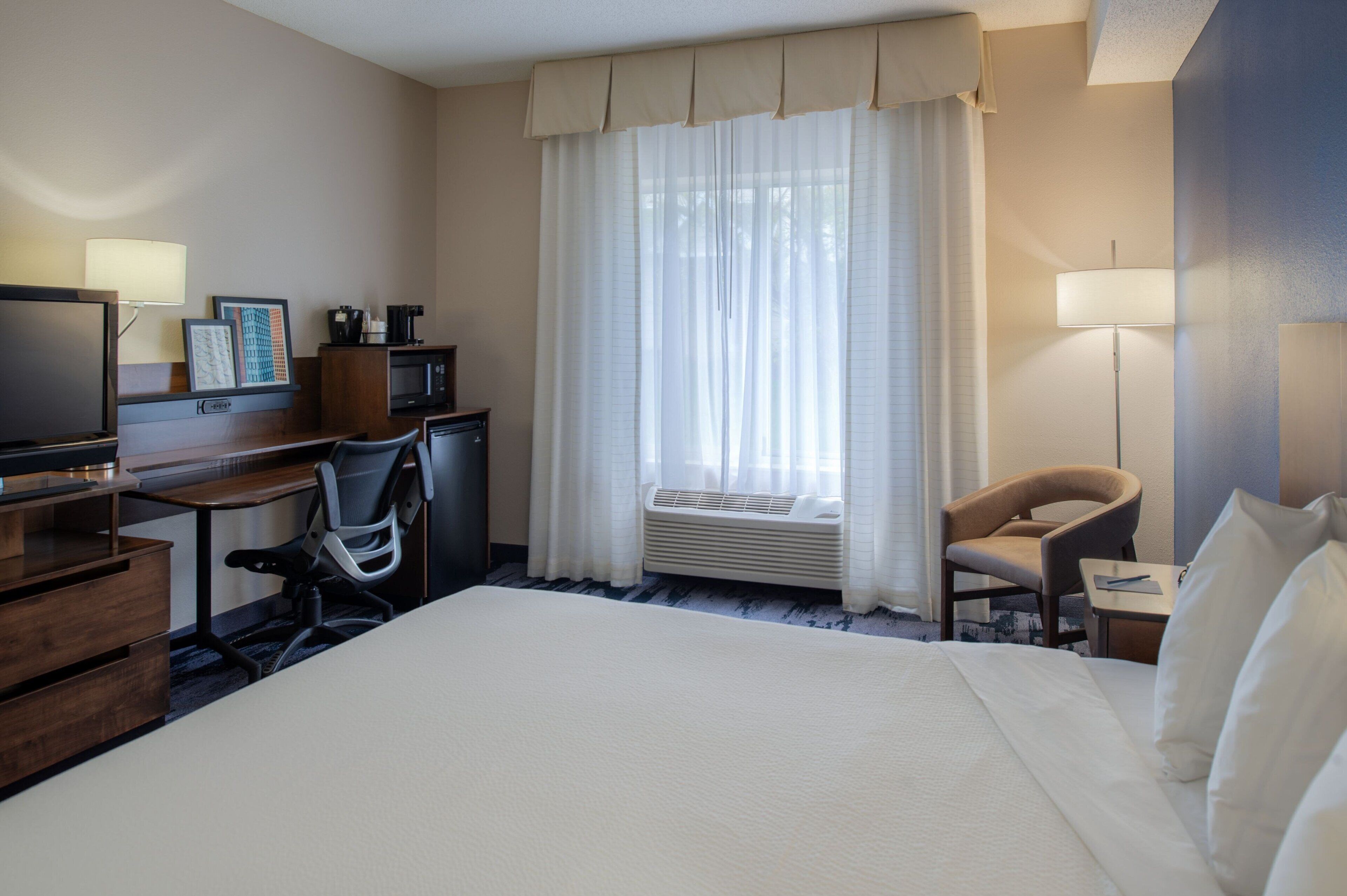 A hotel room with a large bed, a desk with a computer, a chair, a television, and a window with curtains.