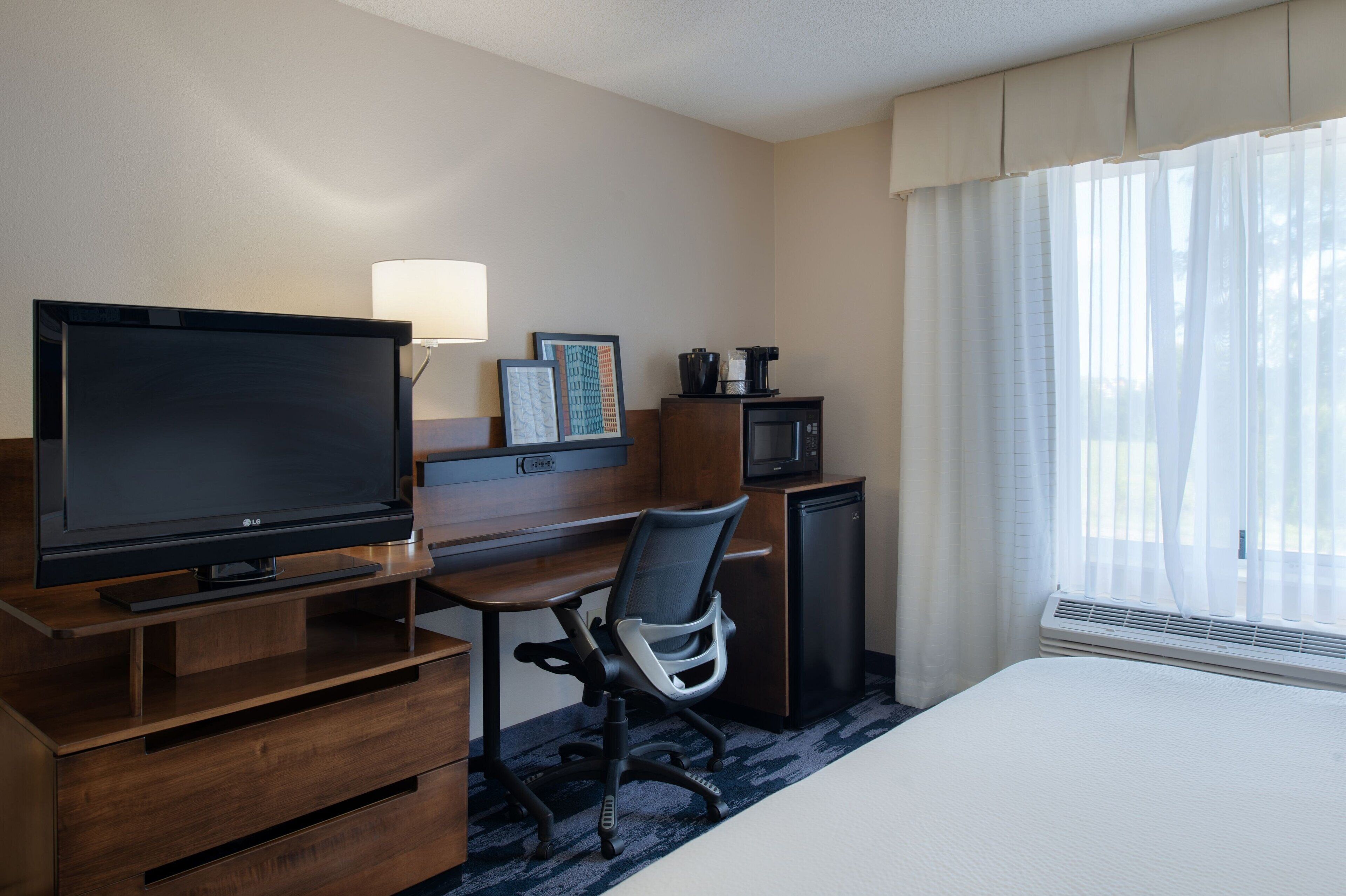 Una habitación de hotel con cama, televisor, escritorio con silla, microondas y ventana con cortinas.