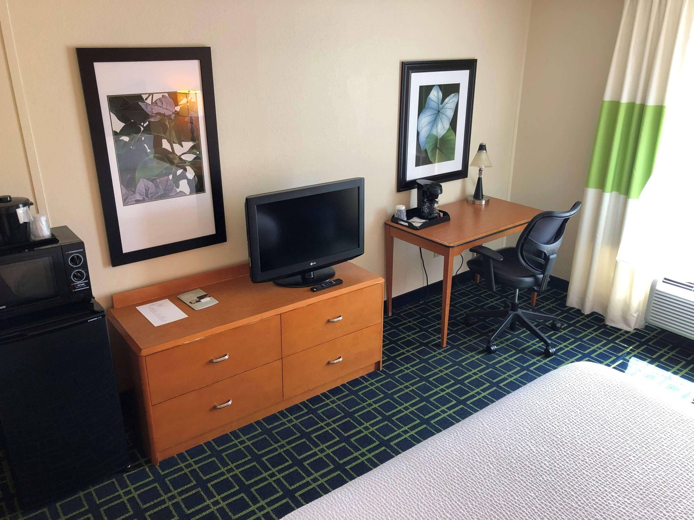 Quarto de hotel com cama, escrivaninha com computador, cadeira, televisão, micro-ondas e cafeteira.