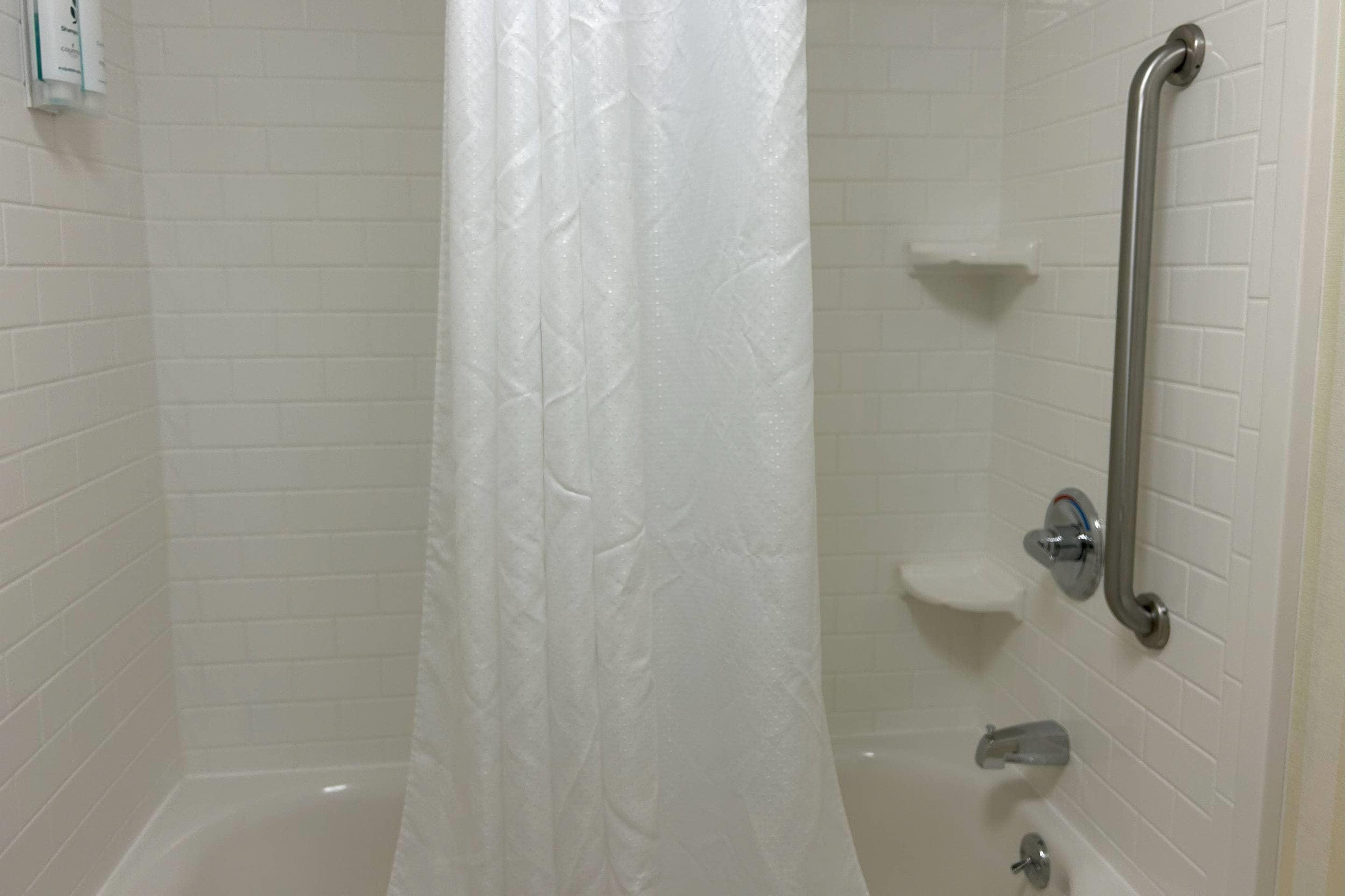 A shower area with a white curtain, a grab bar, and a white tiled wall.