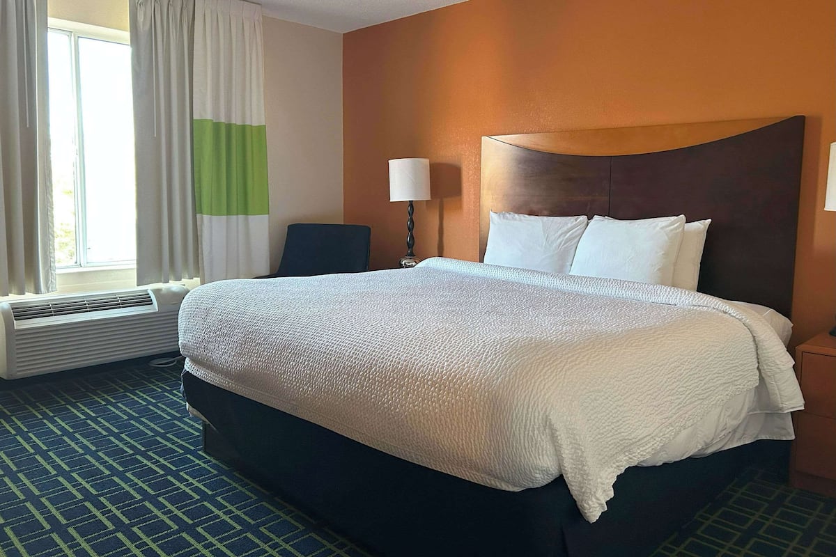 Una habitación de hotel con una cama grande, una silla, una lámpara y un ventanal con cortinas.