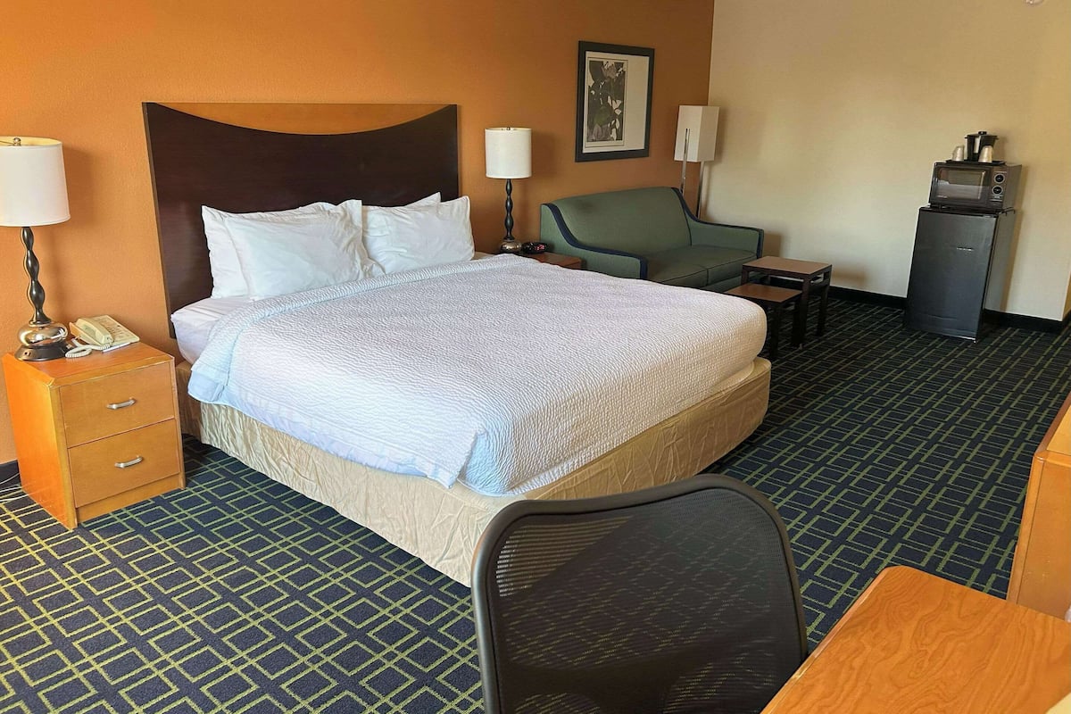 Habitación de hotel con una cama grande, un escritorio, una silla, una mesita pequeña, un refrigerador y un teléfono en la mesita de noche.