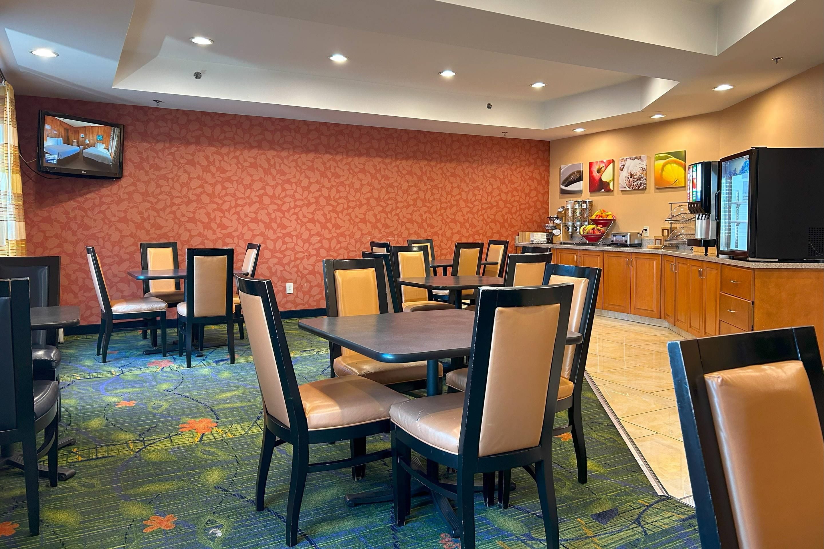 Comedor de hotel con varias mesas y sillas, zona de cocina con refrigerador y cafetera, y un televisor montado en la pared.