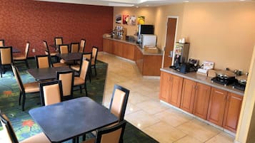 A hotel dining area with multiple tables and chairs, a kitchen counter with appliances, and a patterned carpet.