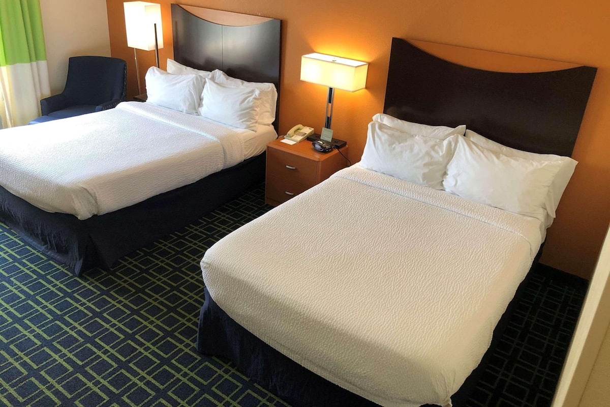 Habitación de hotel con dos camas, una mesita de noche con lámpara, teléfono y una silla.