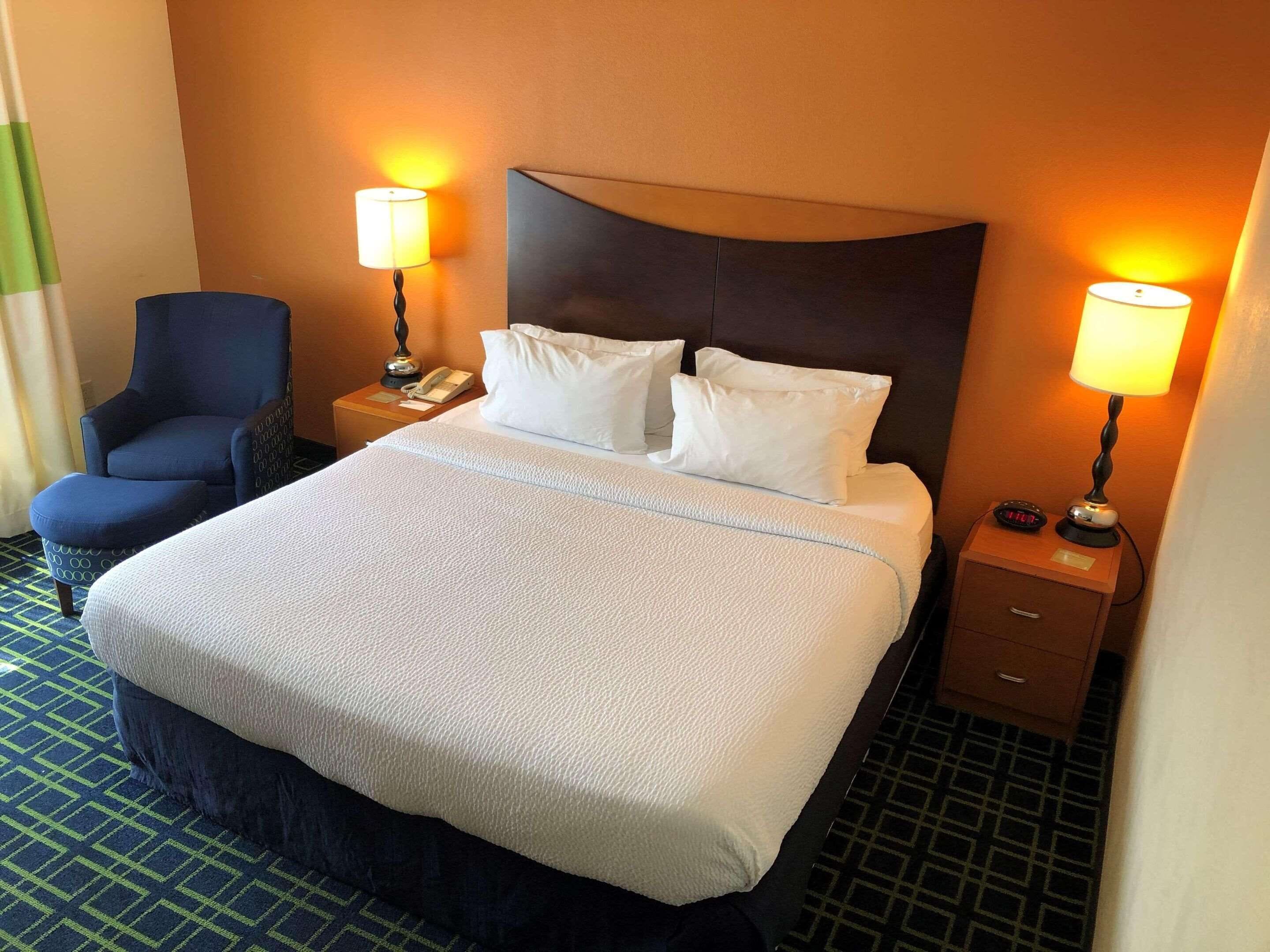 A hotel room with a large bed, two bedside lamps, a chair, a nightstand, and a wall-mounted clock.