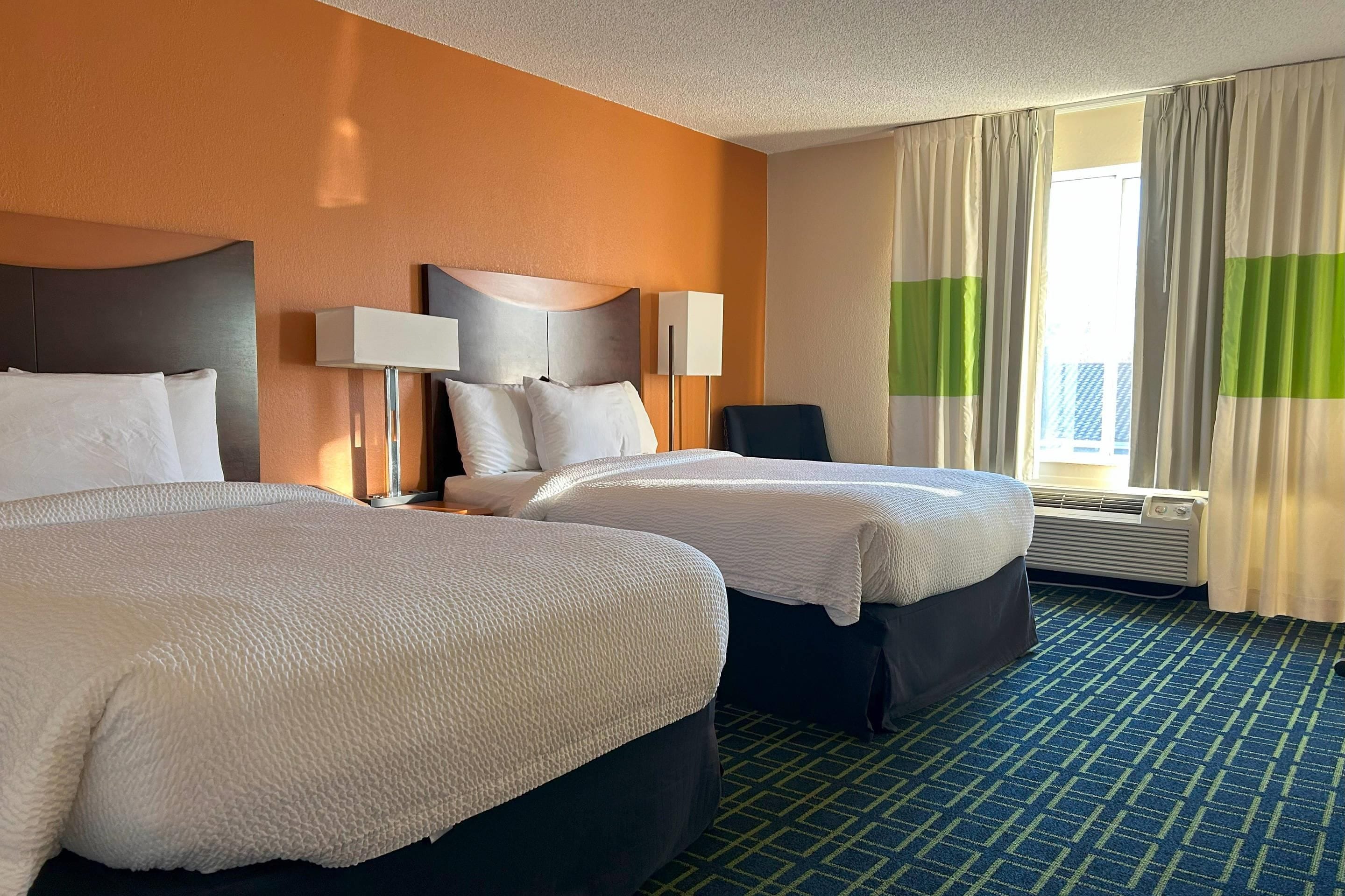 Una habitación de hotel con dos camas, una mesita de noche, una lámpara, una silla y una ventana con cortinas.