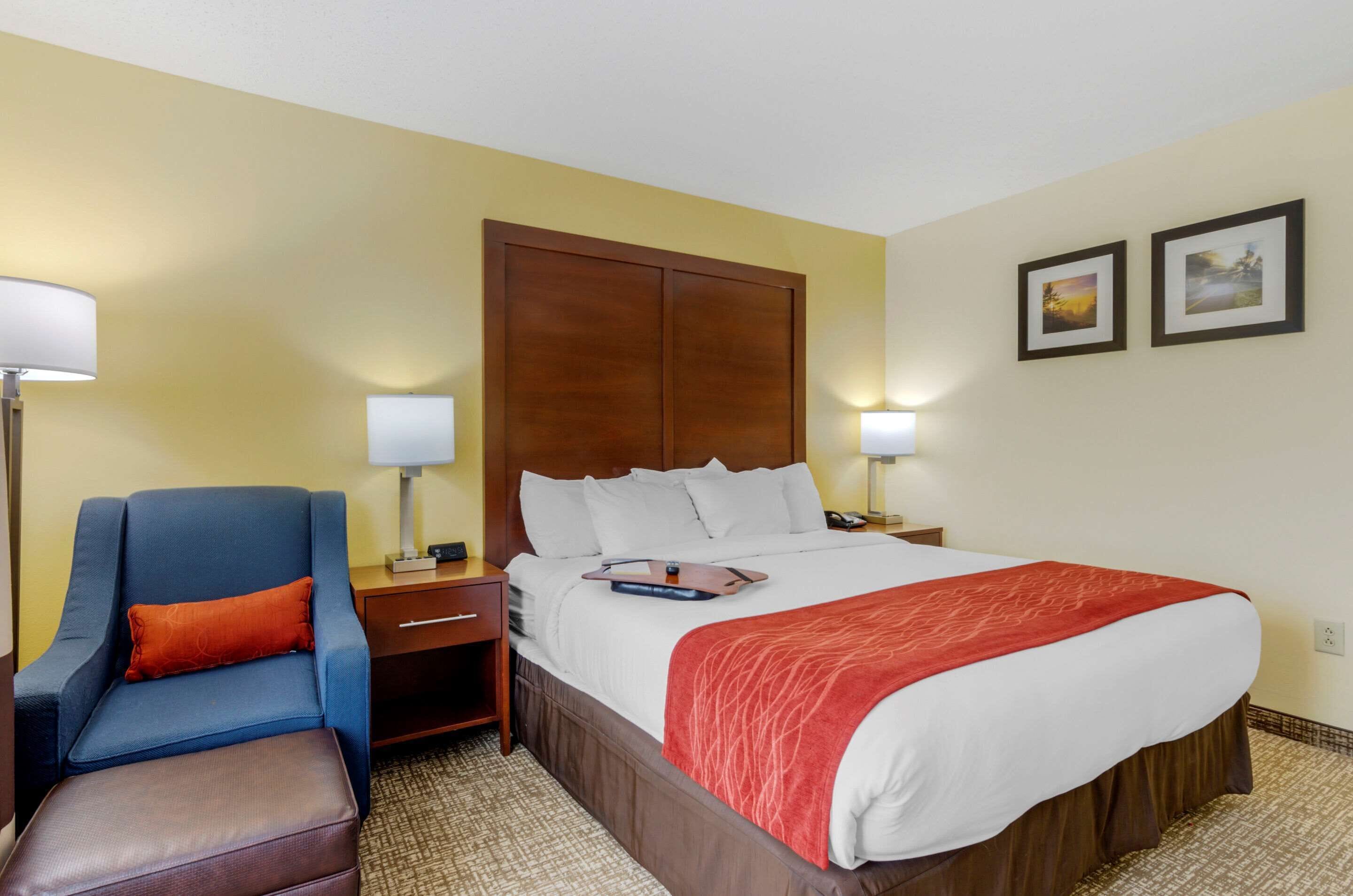 A hotel room with a bed, a blue armchair, a wooden nightstand, a red throw blanket, and two framed pictures on the wall.