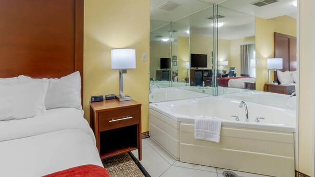 A hotel room with a bed, a bedside table with a lamp, a large mirror, and a bathtub. - Comfort Inn & Suites Hillsville I-77 (Hillsville)