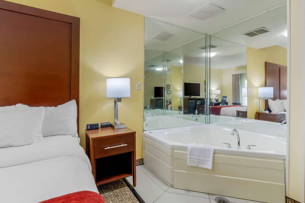 Une chambre d’hôtel avec un lit, une table de chevet avec une lampe, un grand miroir et une baignoire.