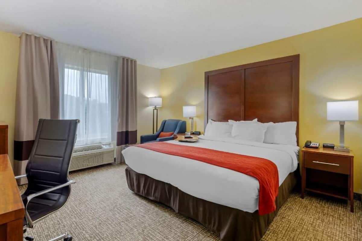 Une chambre d’hôtel comprenant un lit, un bureau, une chaise, une table de chevet et une fenêtre avec des rideaux.