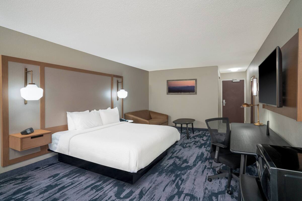 A hotel room with a large bed, a desk with a TV, a chair, a small table, and a nightstand with a phone.