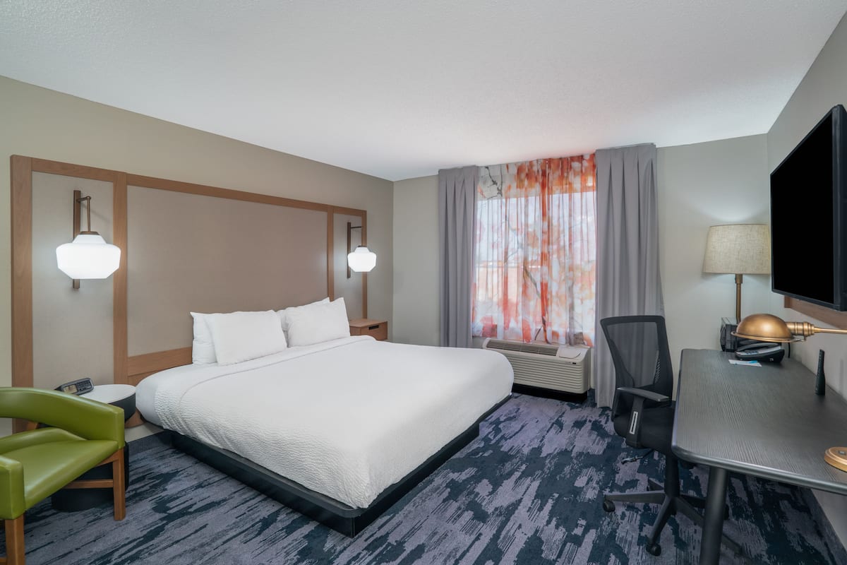 A hotel room with a large bed, a desk with a lamp, a chair, a TV, and a window with curtains.