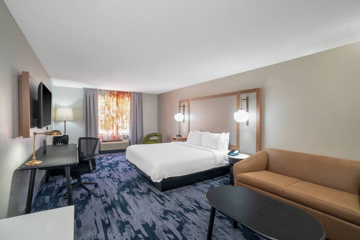 A hotel room with a bed, a desk with a lamp, a sofa, a TV, and a window with curtains.