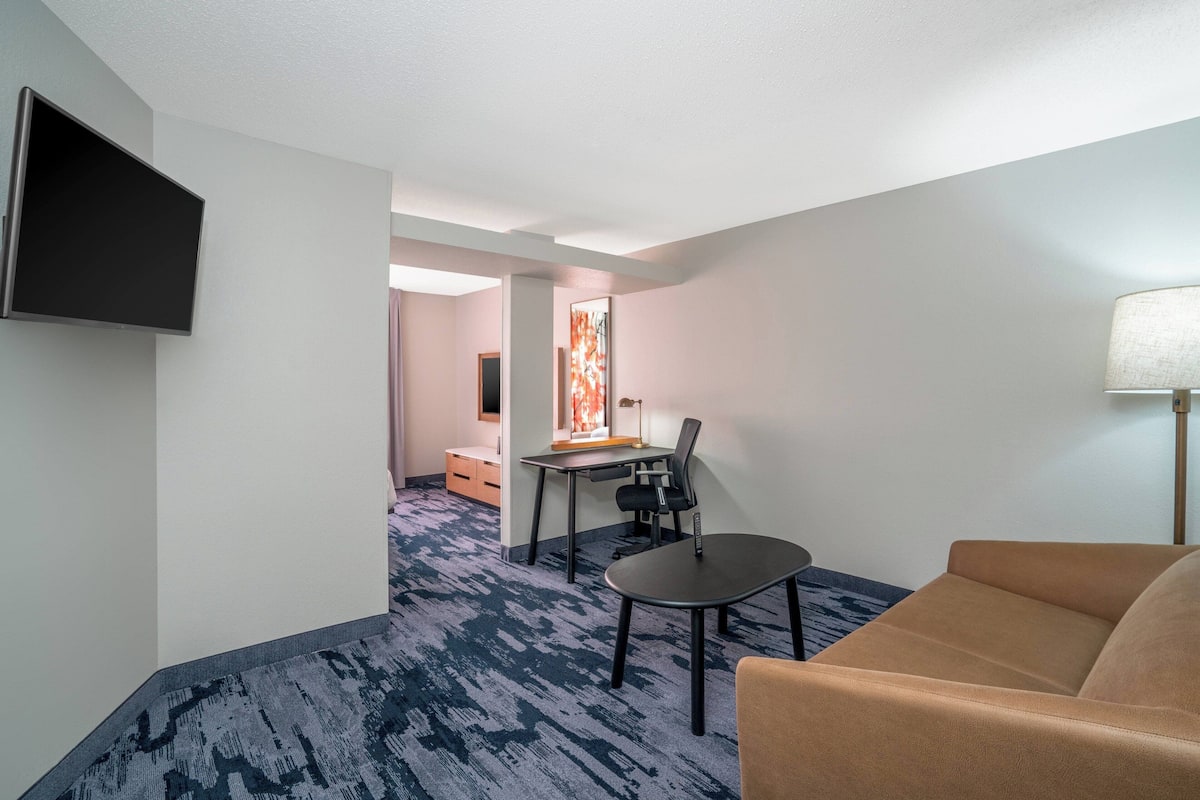 A hotel room with a flat-screen TV mounted on the wall, a desk with a chair, a sofa, and a lamp.