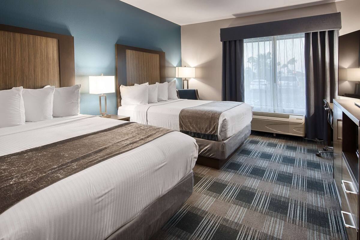 Habitación de hotel con dos camas, un ventanal grande con cortinas, un escritorio y un televisor de pantalla plana, además de un calentador.