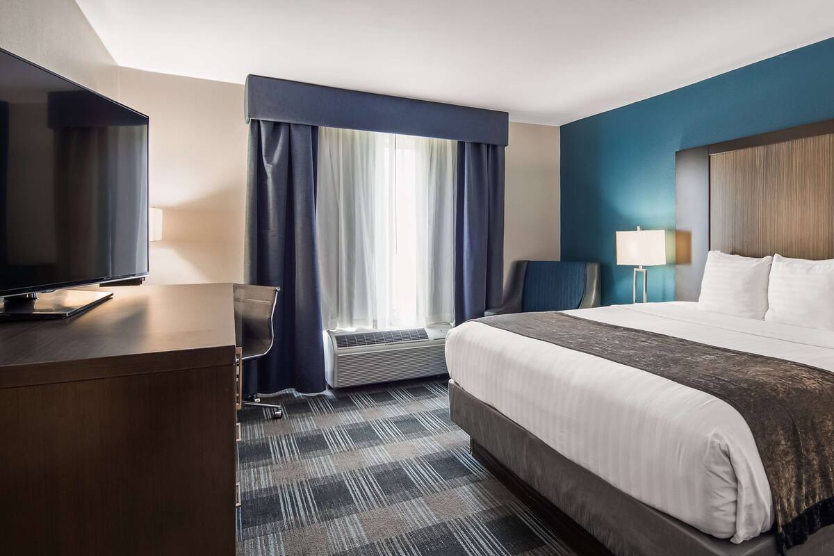 Una habitación de hotel con cama, escritorio, televisor, lámpara y ventana con cortinas.