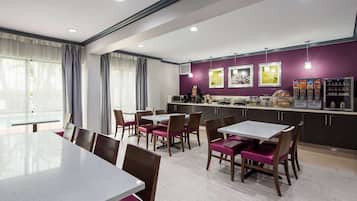 A modern dining area with a long table and chairs, a kitchen counter with appliances and a refrigerator, and a purple accent wall with artwork.