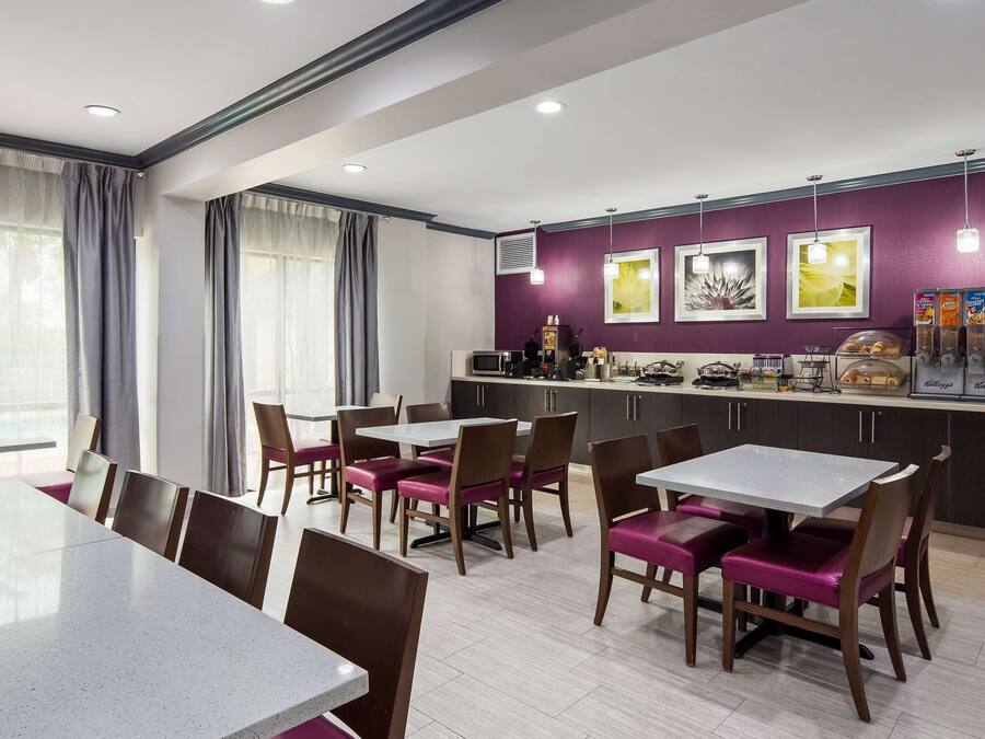 A modern dining area with a long table and chairs, a kitchen counter with appliances and a refrigerator, and a purple accent wall with artwork.