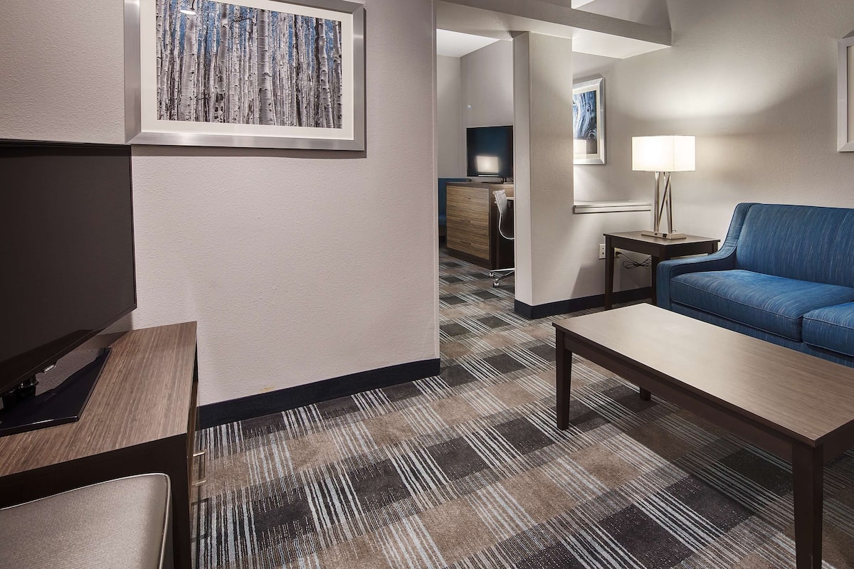 Habitación de hotel con sofá azul, mesa de centro de madera, alfombra estampada y cuadros en las paredes.