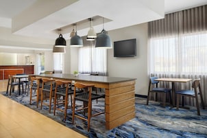 Habitación de hotel con barra de madera, mesa de comedor con sillas, televisor de pantalla plana en la pared y ventanas amplias con cortinas translúcidas.
