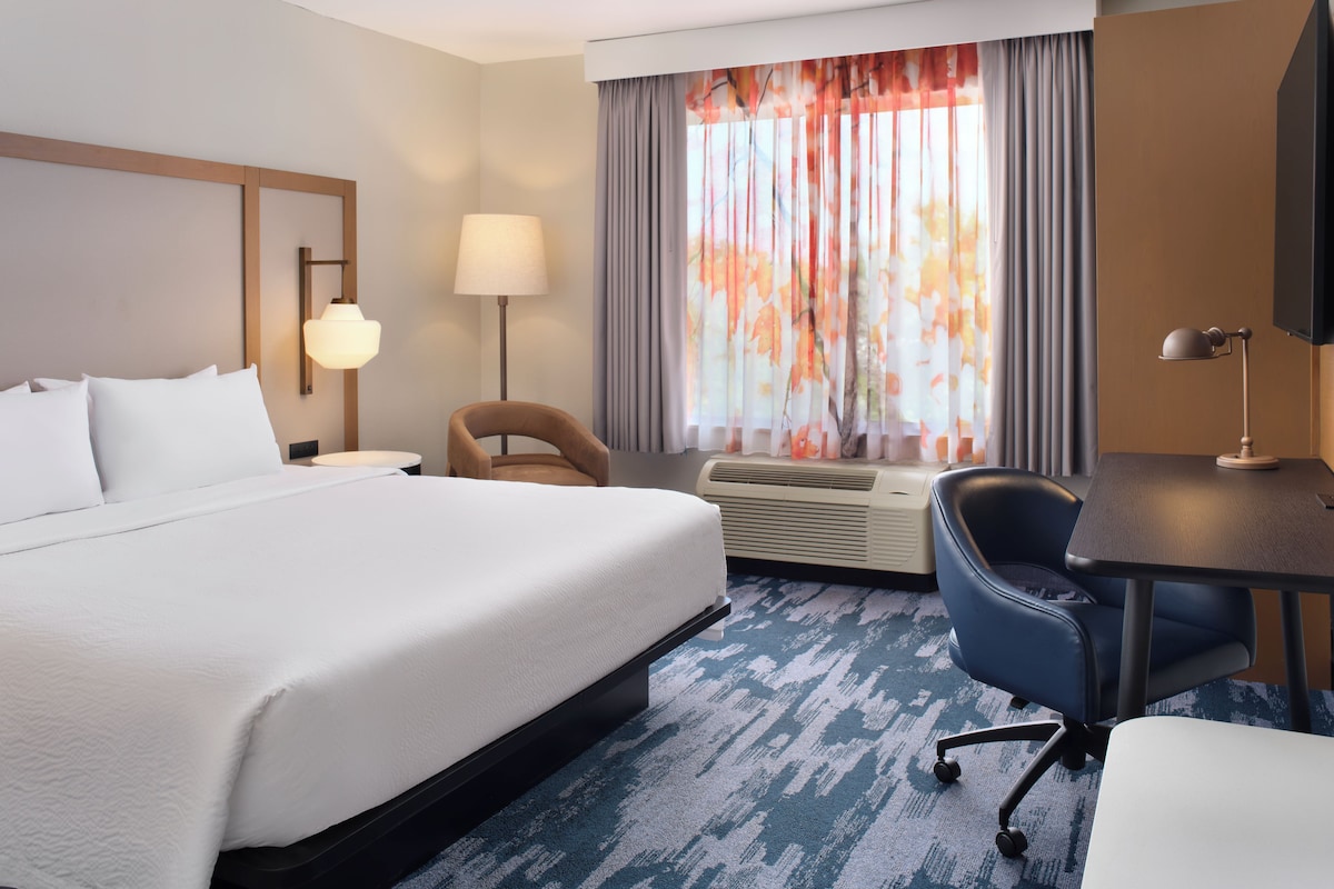 Une chambre d’hôtel avec un lit, un bureau, une chaise, une lampe et une fenêtre avec des rideaux.