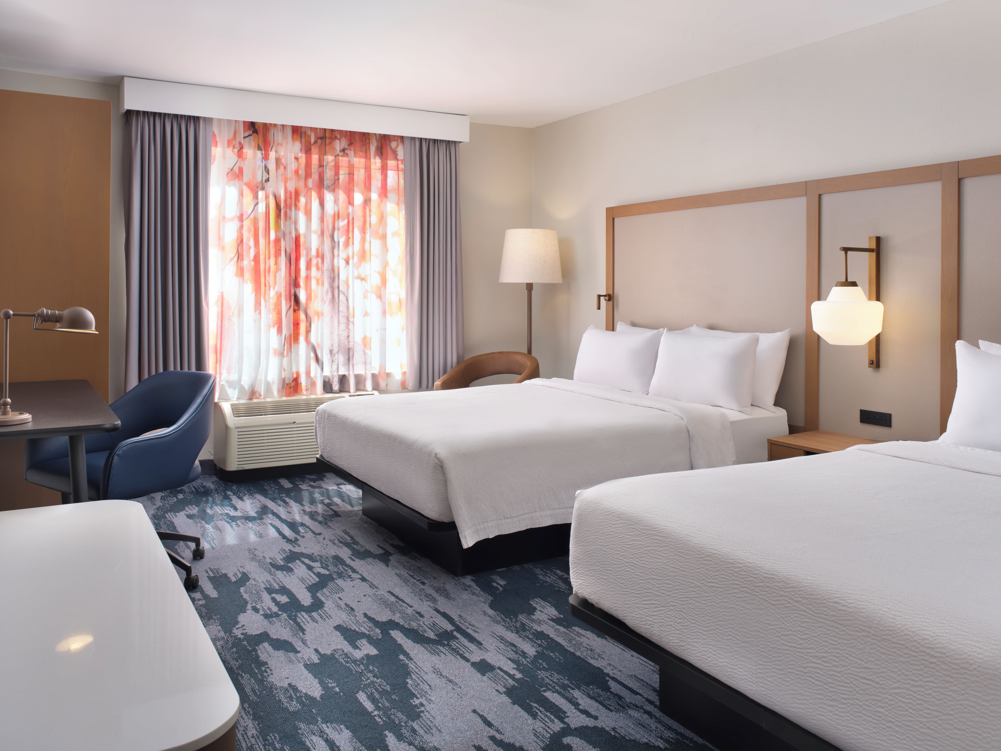 A hotel room with two beds, a desk, a chair, a lamp, and a large window with curtains.