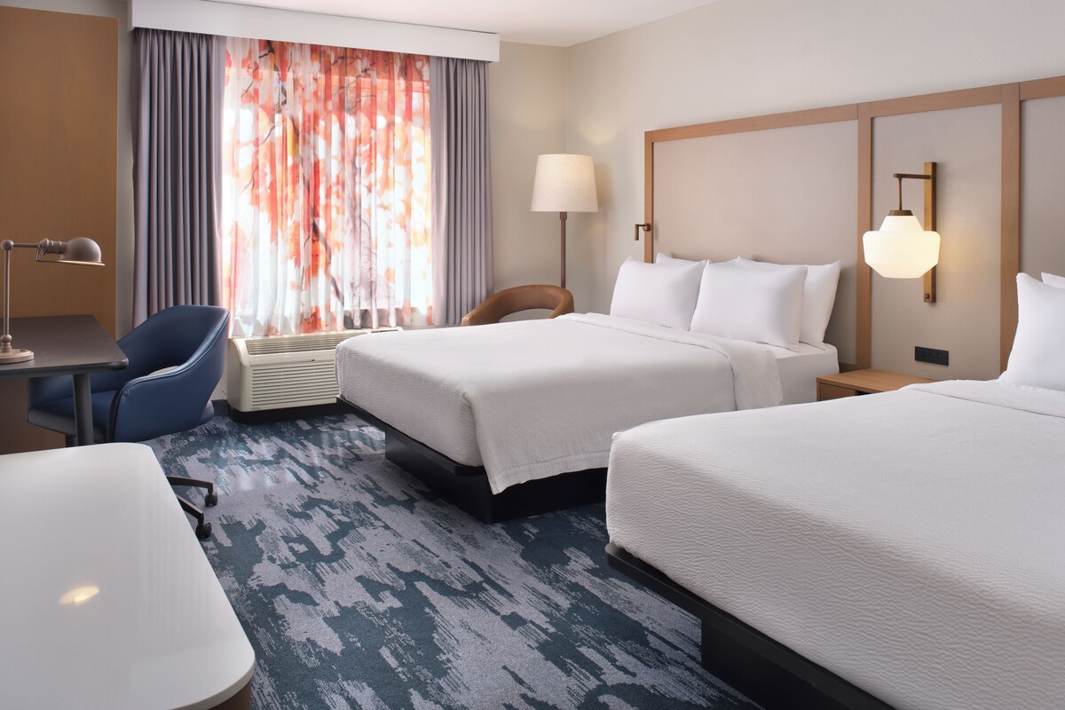 Une chambre d’hôtel avec deux lits, un bureau, une chaise, une lampe et une grande fenêtre avec des rideaux.