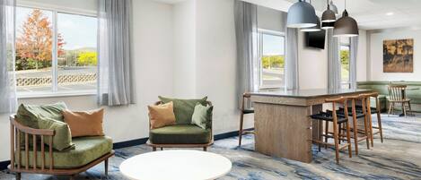 A hotel room with a large window, a green sofa, a round table, and a bar area.