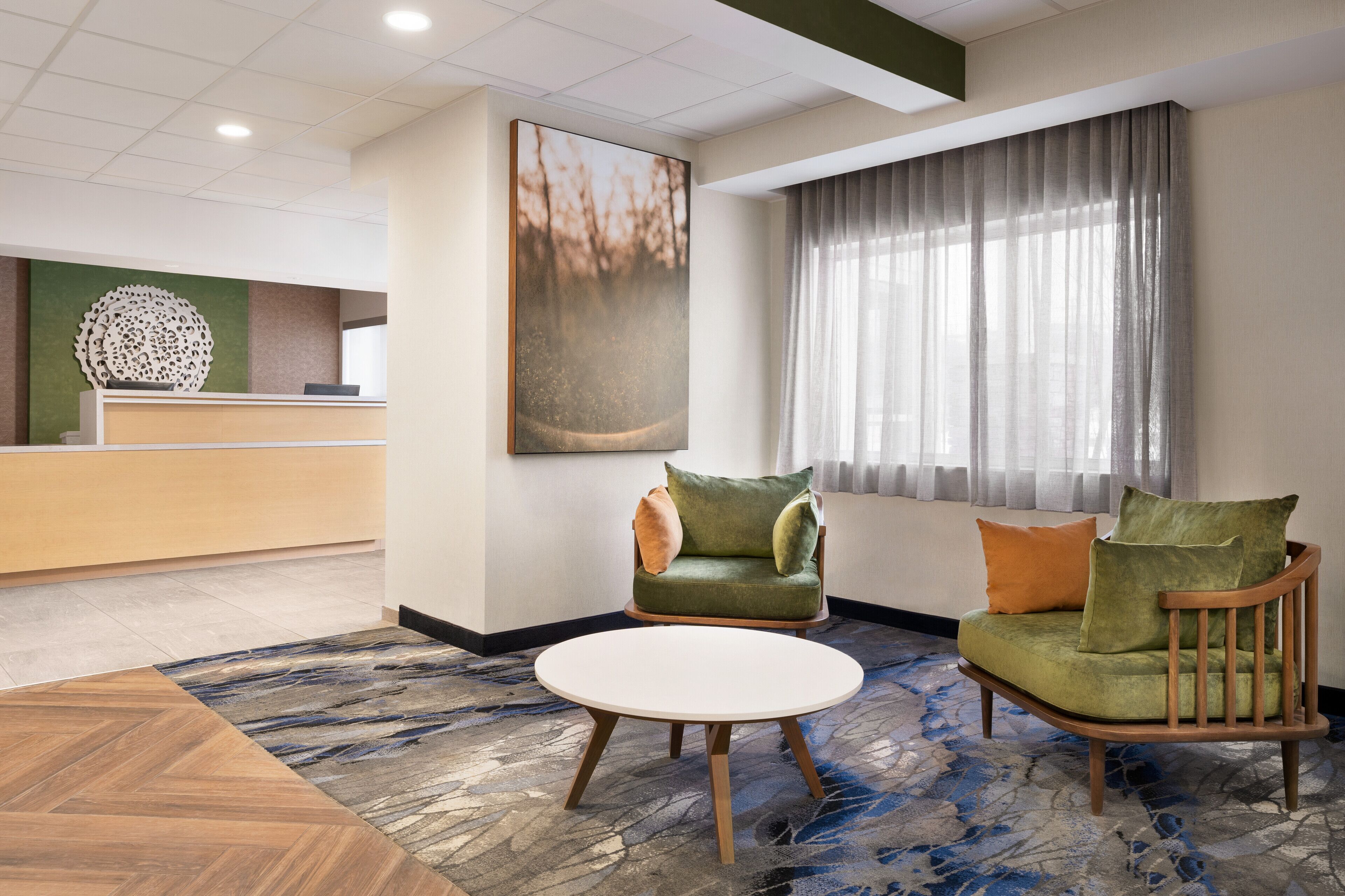 Un hall de hotel con mostrador de recepción, un sofá verde con cojines, una mesa redonda de madera y un cuadro abstracto grande en la pared.