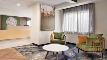 Un hall de hotel con mostrador de recepción, un sofá verde con cojines, una mesa redonda de madera y un cuadro abstracto grande en la pared.