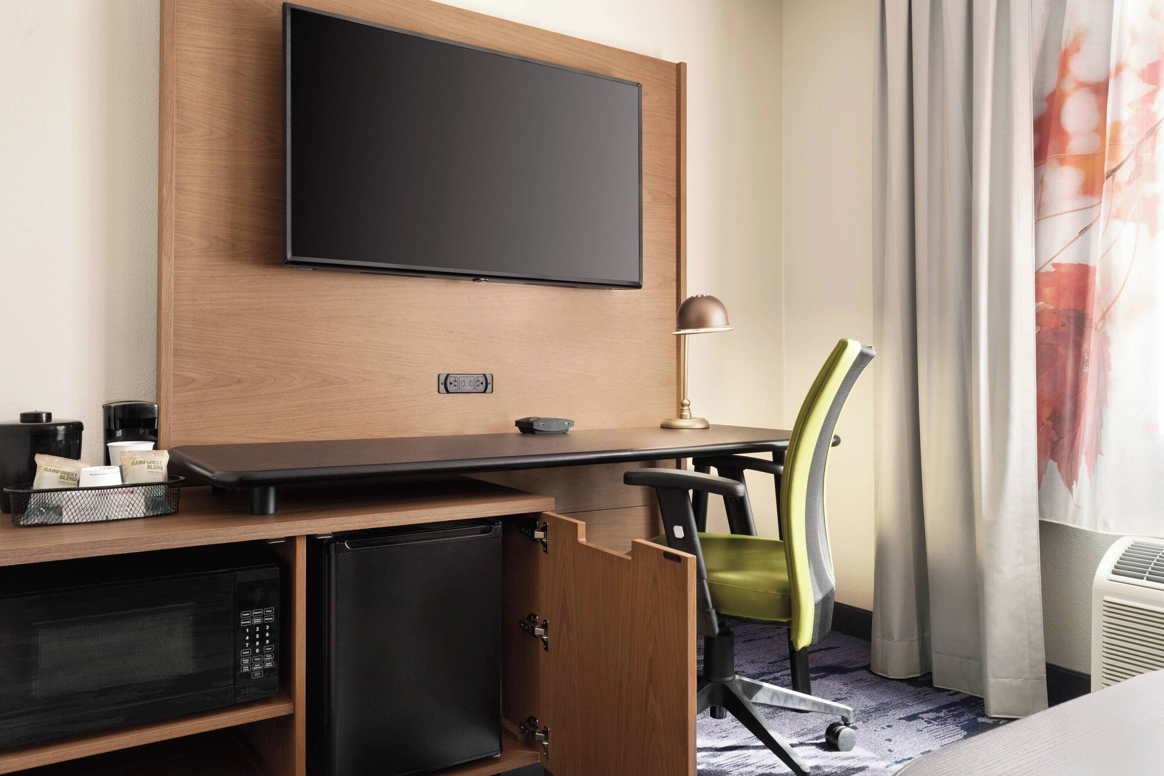 A hotel room with a flat-screen TV mounted on a wooden wall, a desk with a lamp, a microwave, a mini fridge, and a green chair.