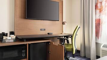 Habitación de hotel con televisión de pantalla plana montada en una pared de madera, un escritorio con lámpara, microondas, refrigerador pequeño y una silla verde.