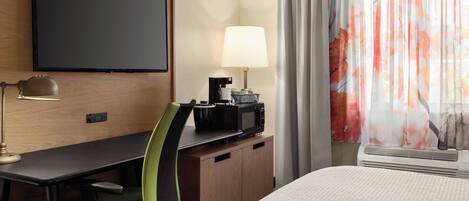 A hotel room with a bed, a desk with a chair, a television, a lamp, a coffee maker, and a window with curtains.