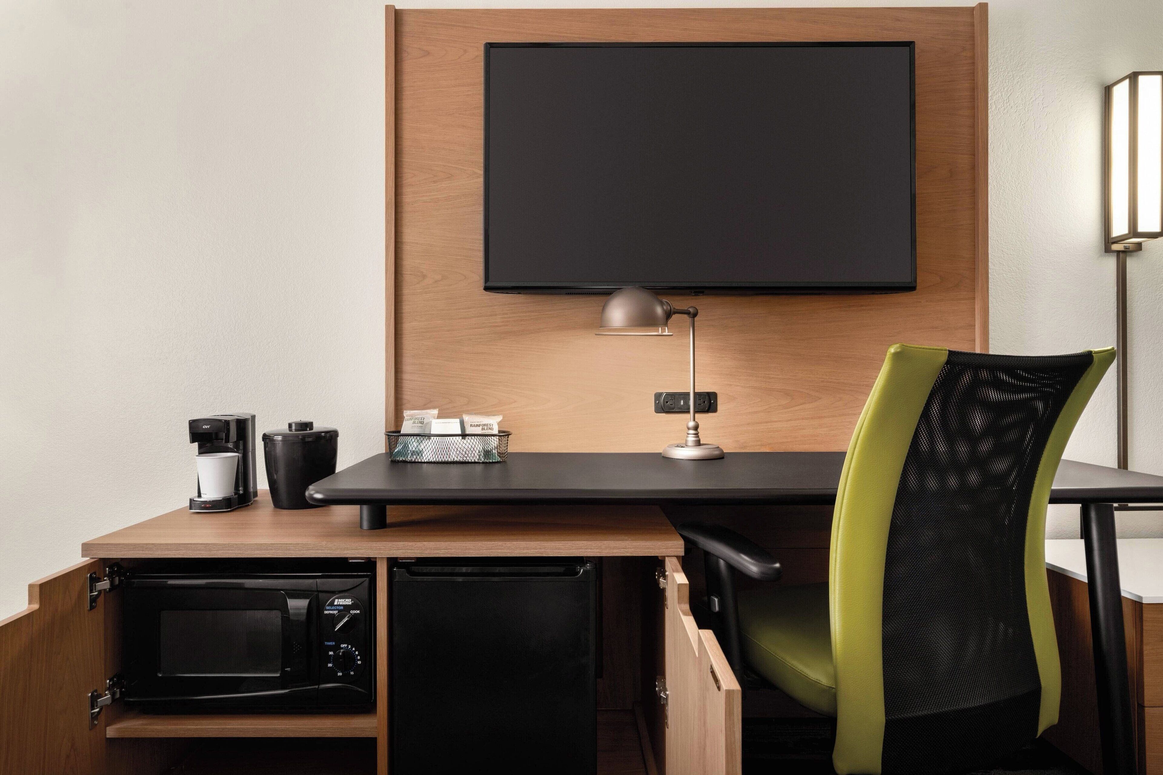 A compact workspace with a flat-screen TV, a desk lamp, a coffee maker, a microwave, and a green mesh chair.