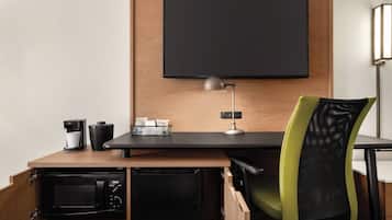 A compact workspace with a flat-screen TV, a desk lamp, a coffee maker, a microwave, and a green mesh chair.