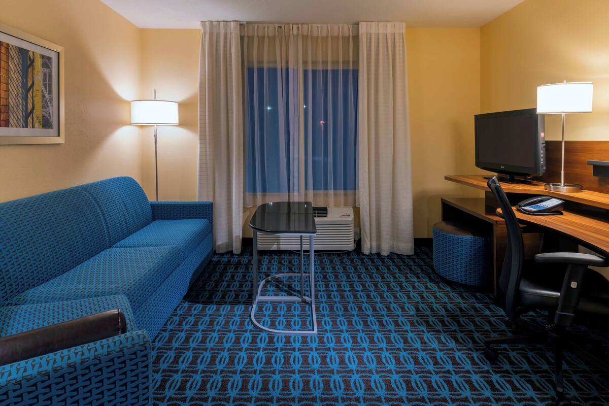 Ein Hotelzimmer mit einem blauen, gemusterten Sofa, einem Schreibtisch mit Computer und Lampe, einem Fenster mit Vorhängen und einem gemusterten Teppich.