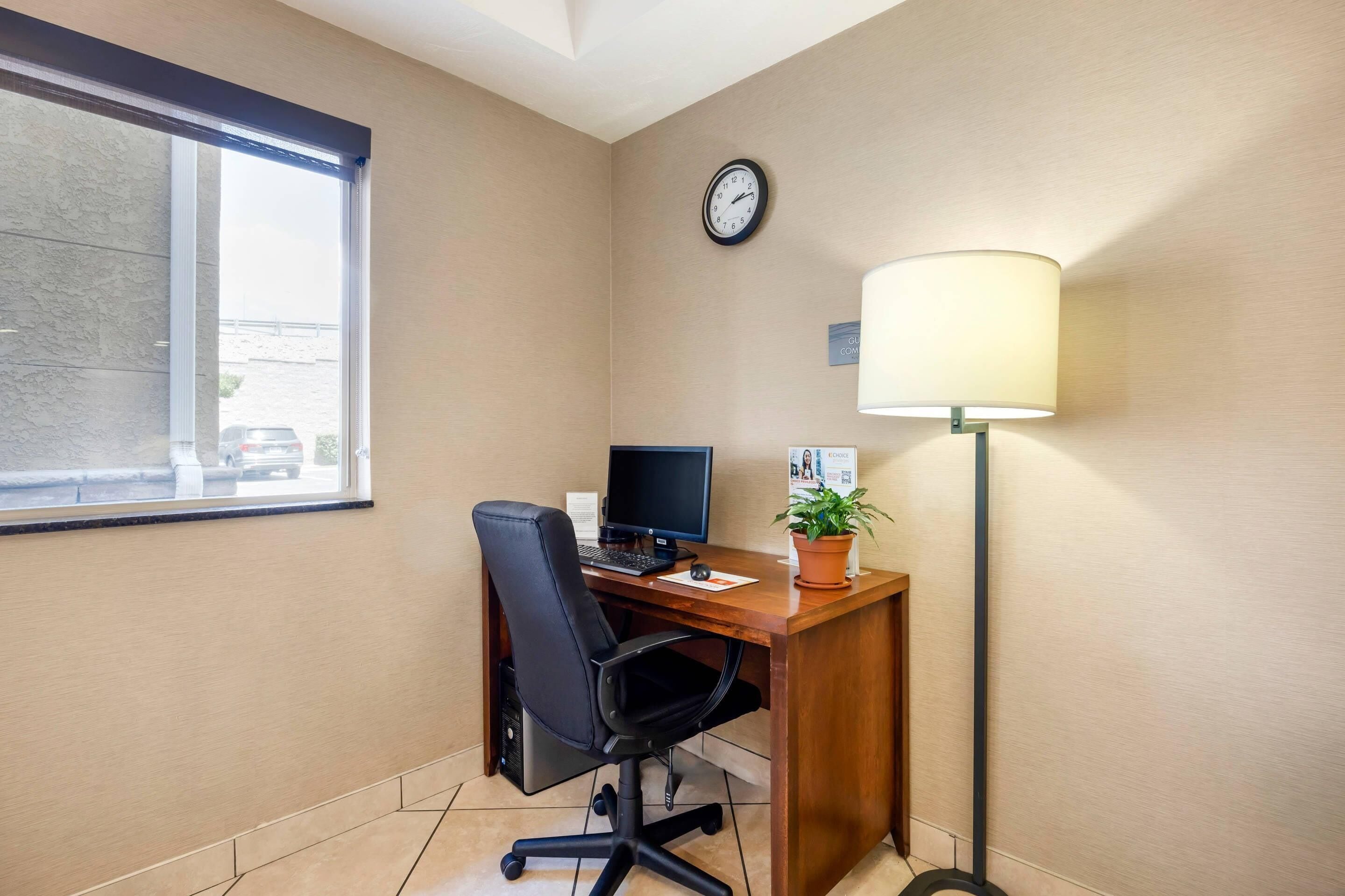A hotel room with a desk, computer, chair, lamp, clock, and window.