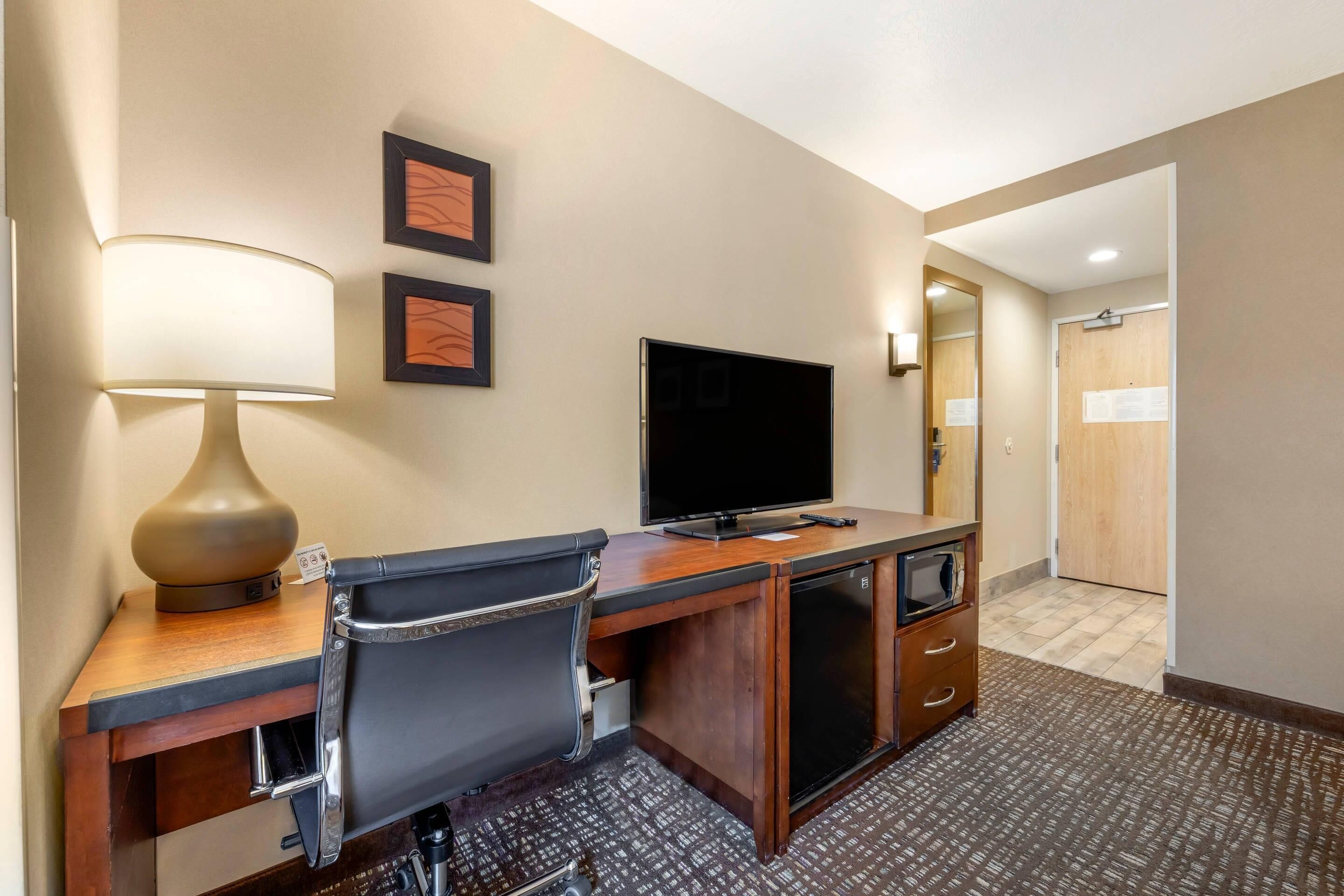 A hotel room with a desk, chair, television, lamp, and a door leading to another room.